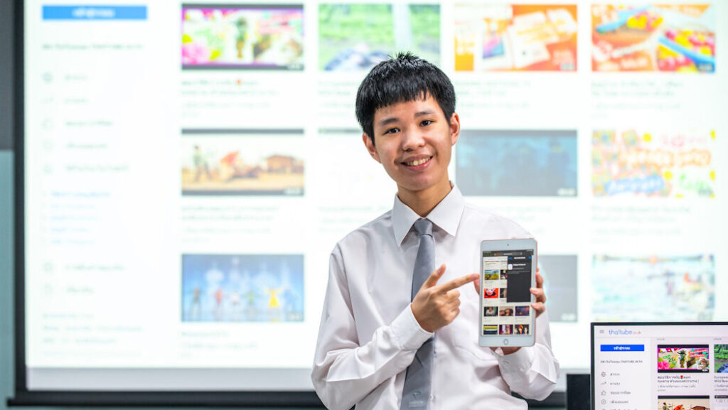 A man in the student uniform inside the computer room. He is seen holding a tablet on one hand with his other hand pointing finger towards the tablet, with his tablet screen opening his former video sharing website Thai Tube..